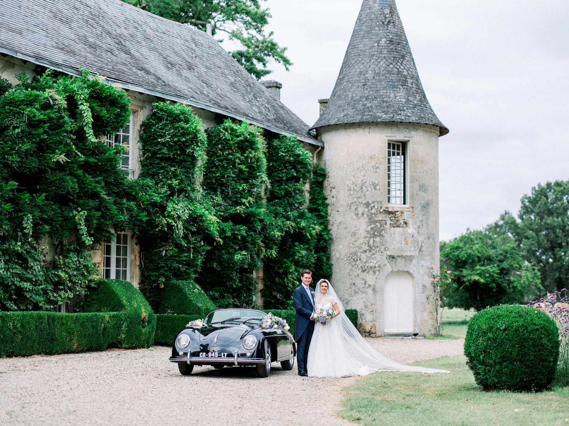 Mariés près d'une voiture ancienne devant un château couvert de lierre à tourelle, en France
